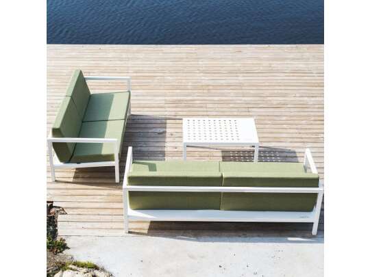 To toseter sofaer i hvit aluminiumsramme og grønne puter, med hvitt bord ute i solen ved vann