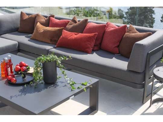 Hjørnesofa i sort aluminium med grå tekstil, pyntet med puter i flere farger og hagebord på terrasse i sollys