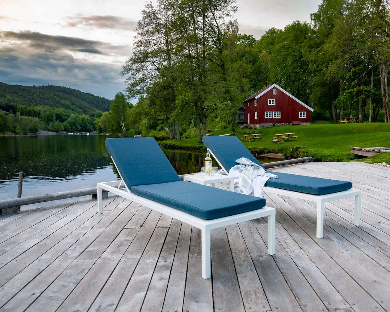 Sundays design solseng i hvit aluminium og blå pute ved vann og hytte